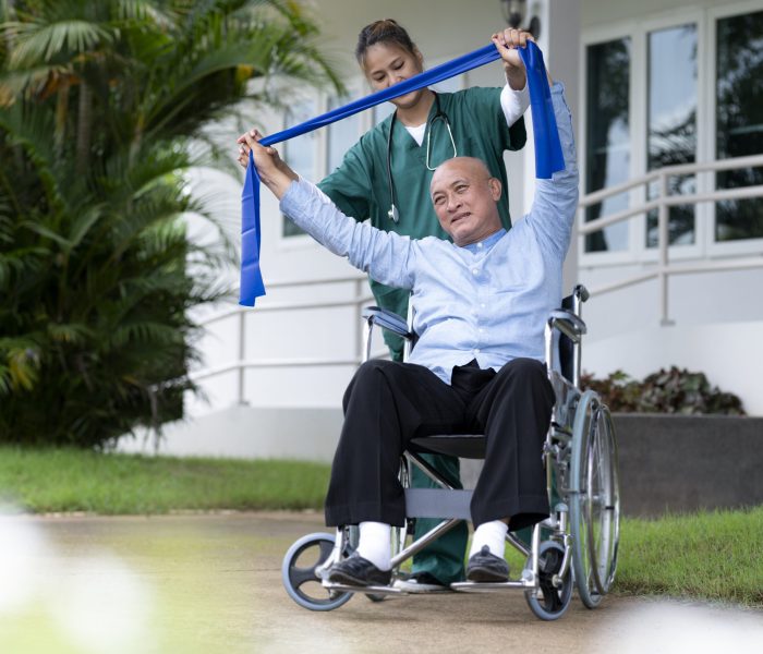 A physiotherapist Asian woman in green clothes helps an elderly man exercise on a wheelchair in a nursing home, nursing care.