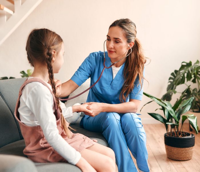 A woman pediatrician doctor at home in a medical uniform with a stethoscope examining a child while sitting on the sofa in the living room. Health care and medicine.